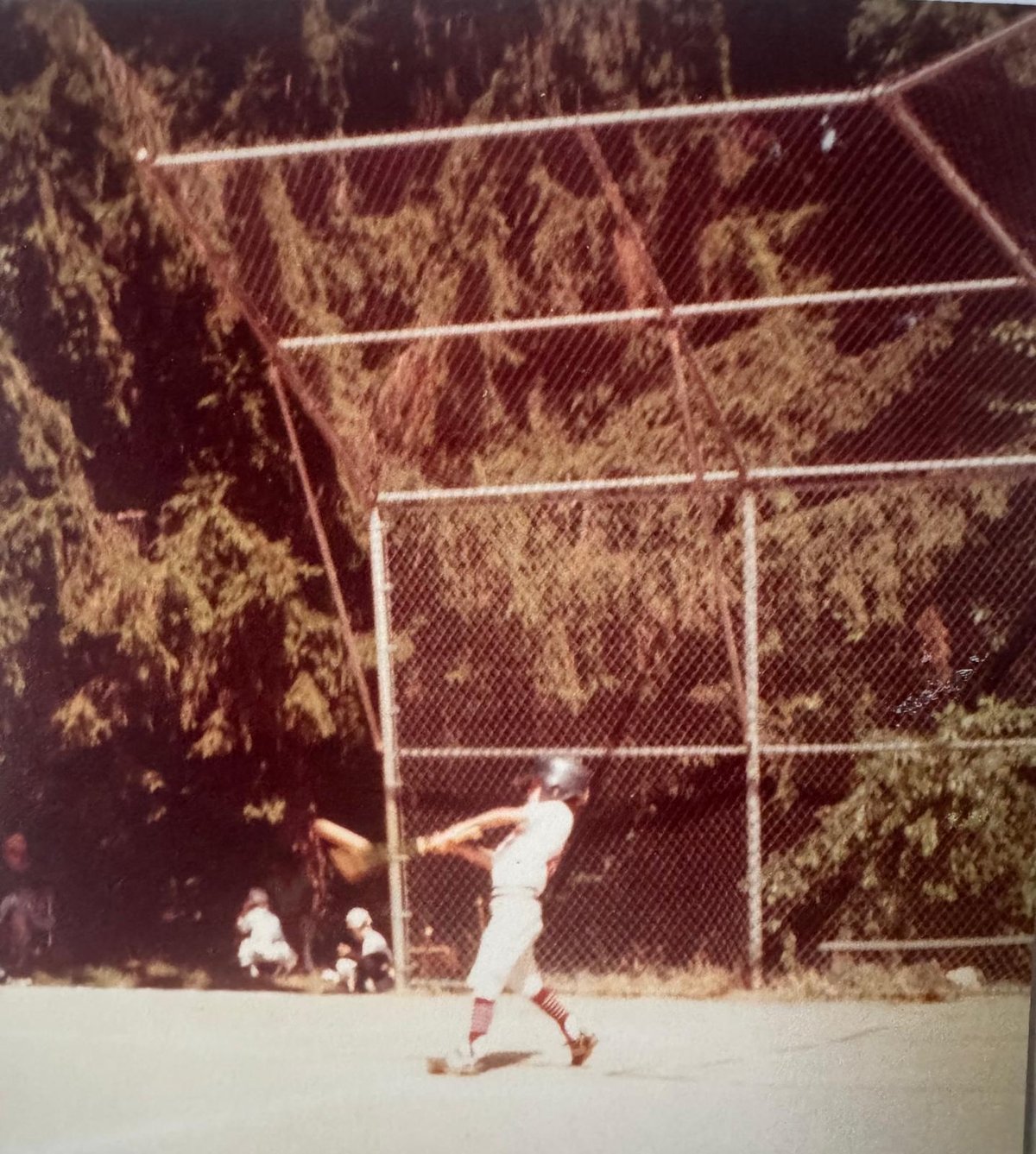 A faded childhood snapshot of David Smith in a Little League uniform, mid-swing at the plate, with a chain-link backstop and tall trees behind him.