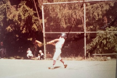 A faded childhood snapshot of David Smith in a Little League uniform, mid-swing at the plate, with a chain-link backstop and tall trees behind him.